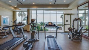 Ein Fitnessstudio mit Laufbändern, Crosstrainern und Kraftgeräten, große Fenster mit Blick auf Palmen und das Meer.