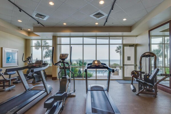 A gym with treadmills, elliptical machines, and weight equipment, large windows with a view of palm trees and the ocean. - Aqua Beach Resort by Panhandle Getaways (Panama City Beach)