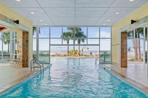 Une piscine intérieure avec des parois en verre transparent, un sol carrelé et une vue sur les palmiers à l’extérieur.