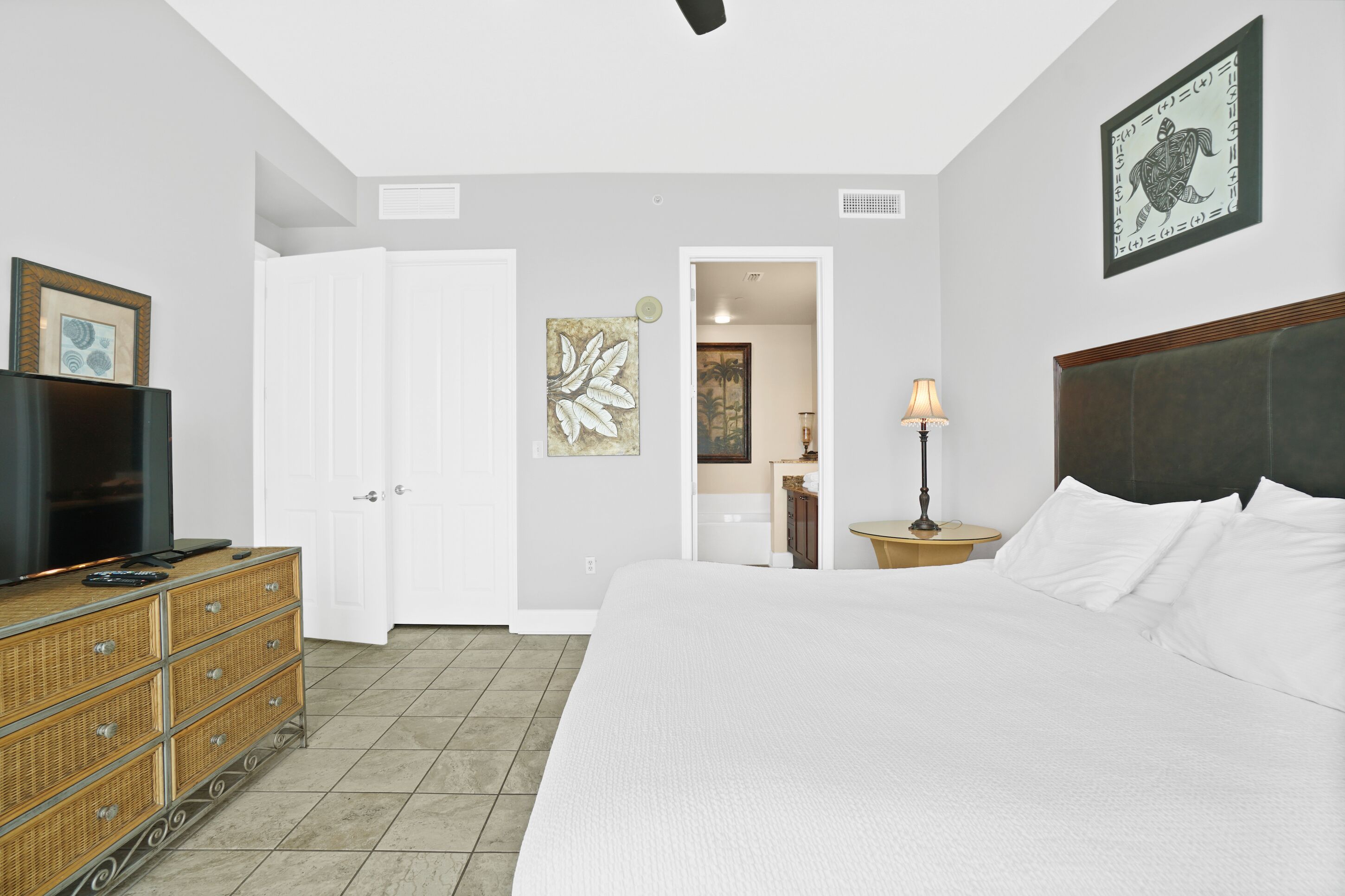 A hotel room with a large bed, a television on a dresser, a door leading to a bathroom, and a framed artwork on the wall.
