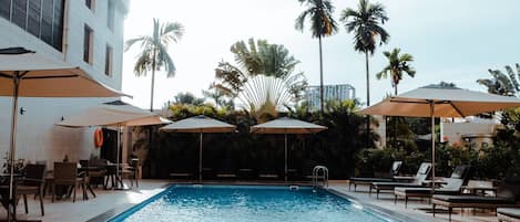 A swimming pool with lounge chairs and umbrellas, surrounded by palm trees and a building.
