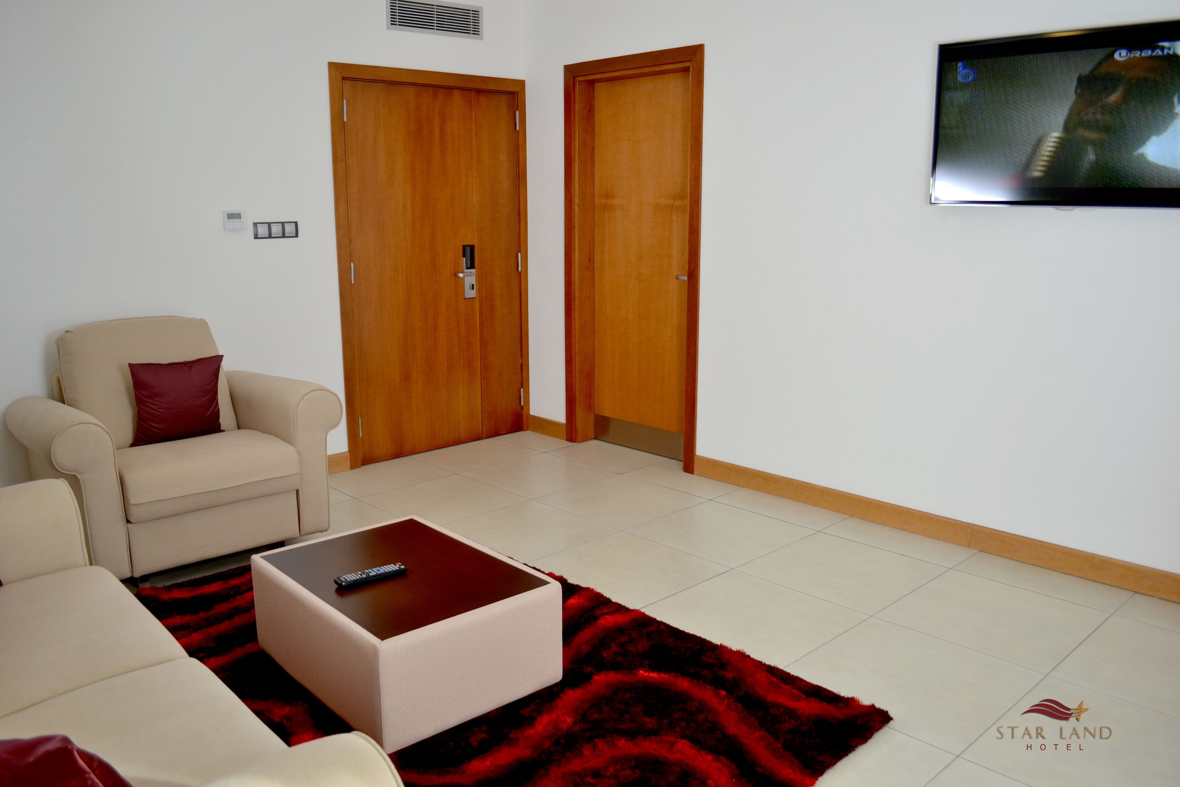 Habitación de hotel con un sofá color beige, una alfombra roja, una mesa de centro de madera y un televisor montado en la pared.