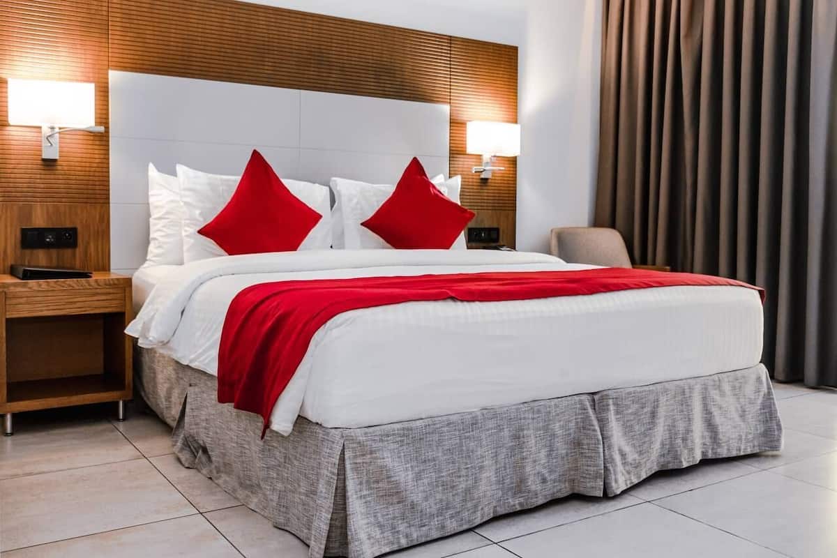 A hotel room with a large bed, two red pillows, a white comforter, a grey skirted bed base, wooden headboard, two bedside lamps, a nightstand, and a chair.