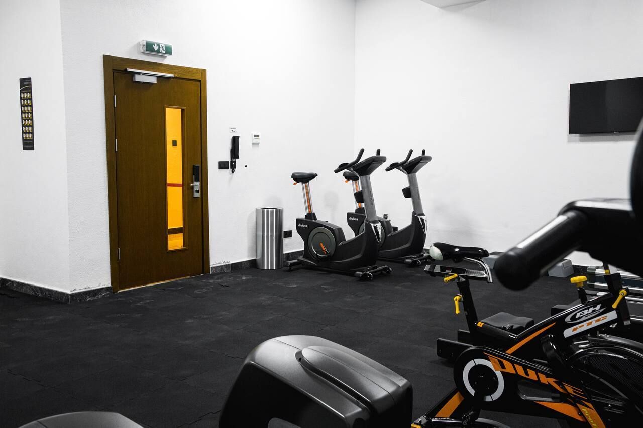 Een fitnessruimte met hometrainers, loopbanden en een deur met een nooduitgang.'
