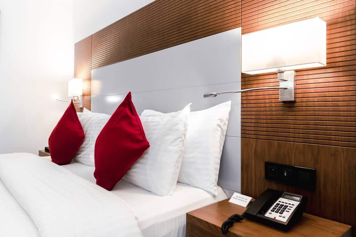 A hotel room with a wooden headboard, white bedding, and red pillows. There is a bedside table with a phone and a lamp.