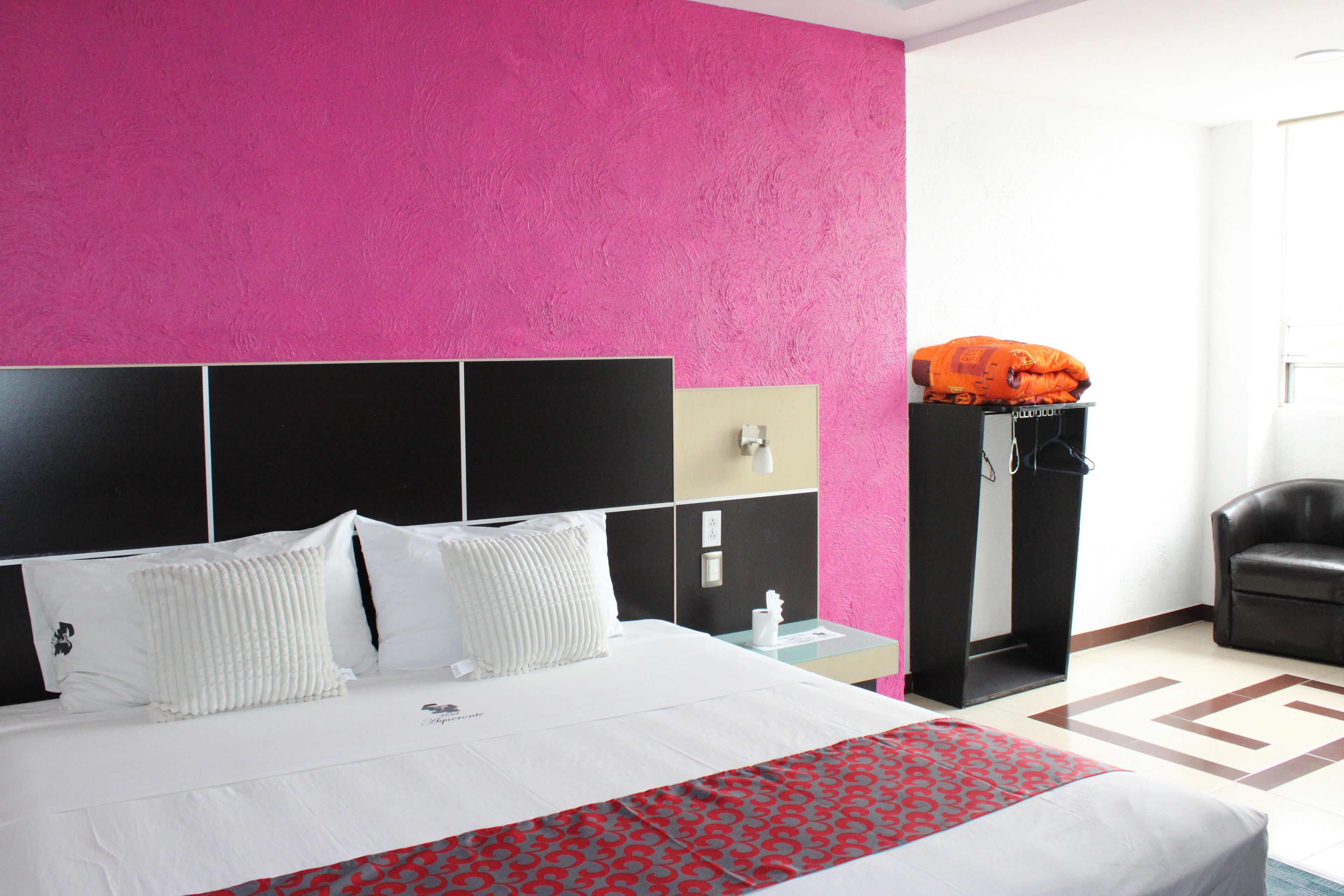 Habitación de hotel con una cama grande, una pared de acento rosa, cabecera negra y una silla de cuero negro.
