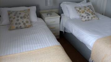 A bedroom with two beds, each with a white duvet and matching pillows. There is a bedside table with a lamp between the beds.