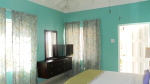 A bedroom with teal walls, a bed with a white and green bedspread, a dresser with a mirror and a flat-screen TV, and floral curtains. - Paradise on the Rocks (Negril)