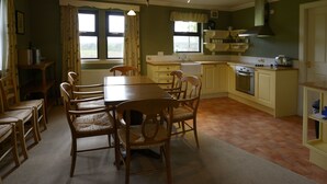 A dining room with a wooden table and chairs, a window with curtains, a kitchen with a stove and sink, and a shelf with items. - Factors Inn & Cottage (Fort William)