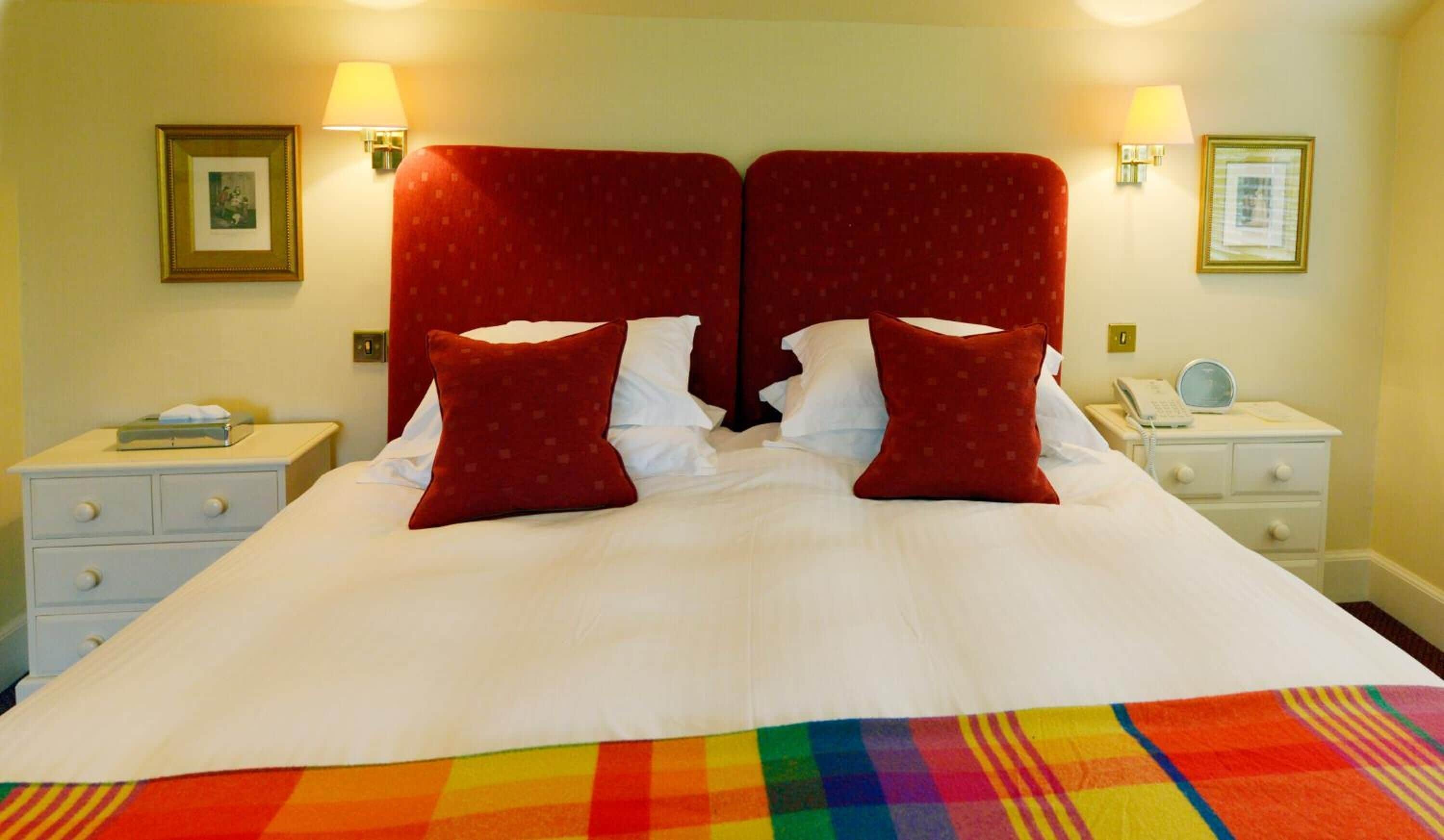 A double bed with red headboards, white bedding, and a colorful blanket. There are two bedside tables with lamps and a clock.