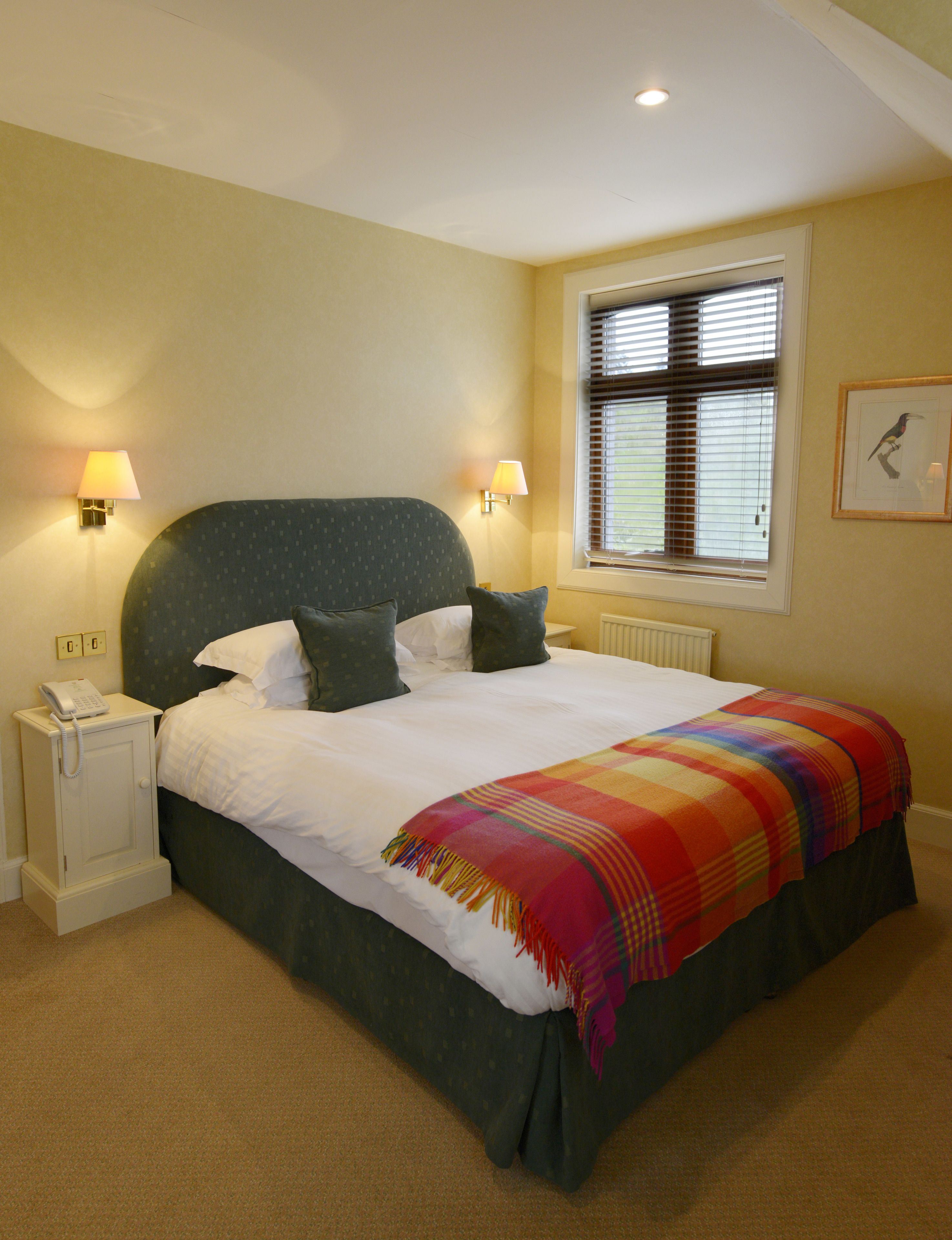 A neatly made bed with a colorful blanket, two bedside lamps, a nightstand with a phone, and a window with blinds.