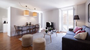 Een moderne woonkamer met een grijze bank, een houten eettafel en een televisie. Er zijn poefs, een bijzettafeltje en een groot raam met uitzicht.
