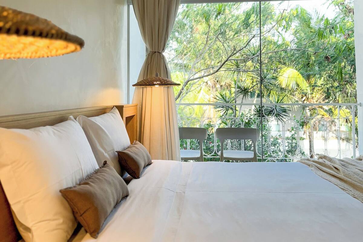 A bedroom with a large bed, a window with curtains, and a view of greenery outside.