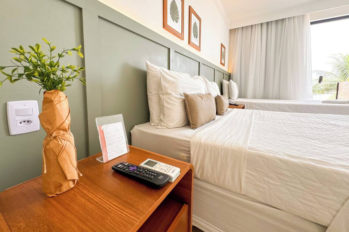 A hotel room with a bed, bedside table, remote control, a plant, and a window with curtains.