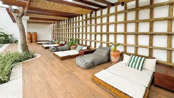 Een moderne buitenlounge met houten vloeren, diverse zitplaatsen zoals banken en bänquettes, en een wand van bamboe.