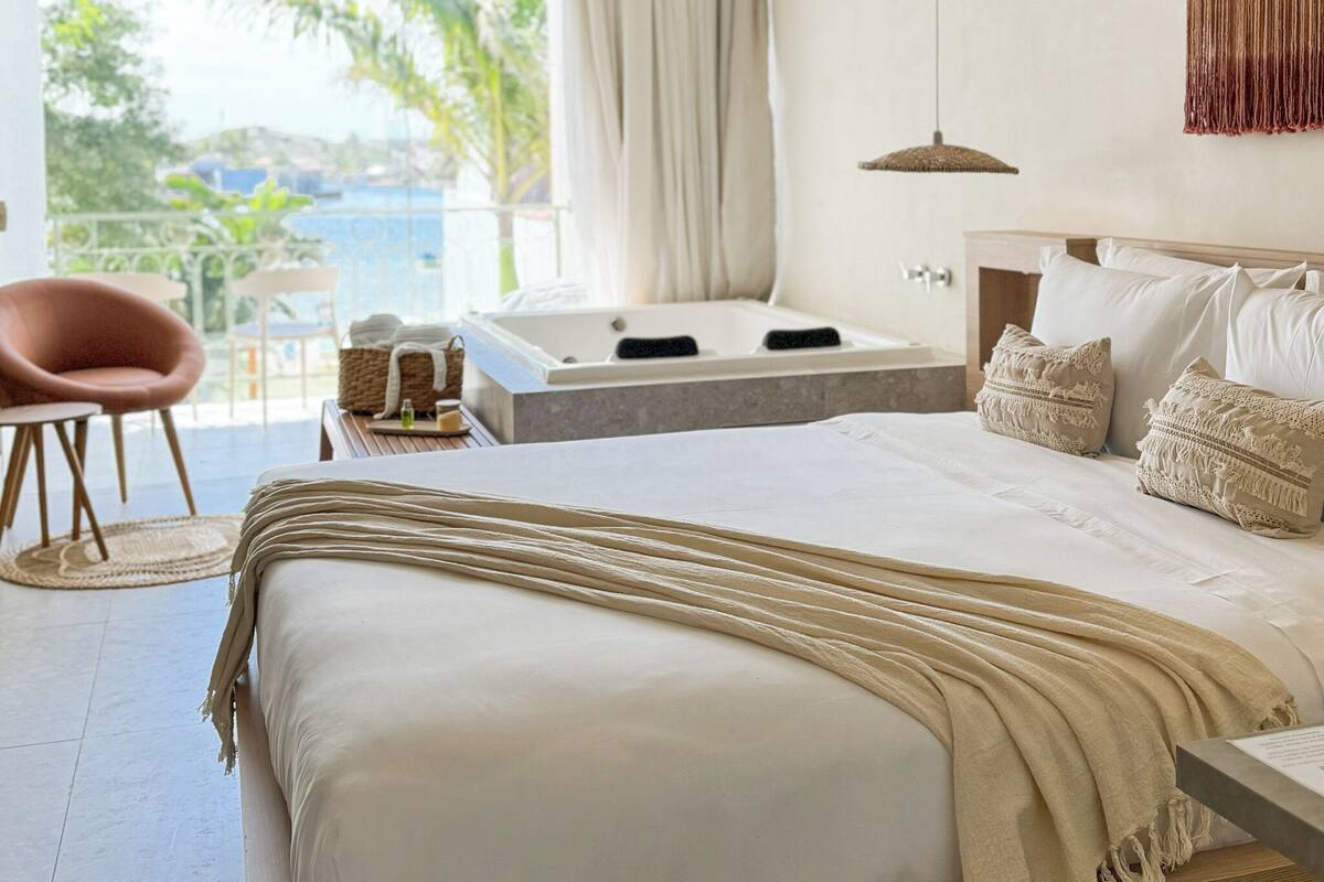 A bedroom with a large bed, a view of the sea, a bathtub, and a chair.