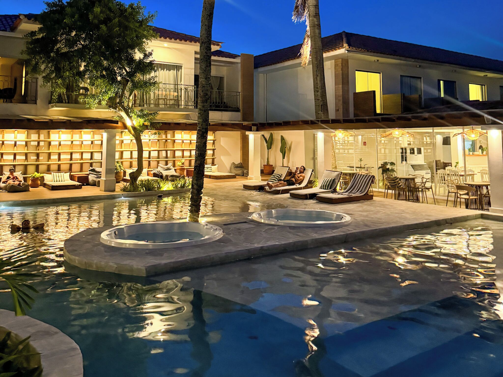 A pool area with lounge chairs and a hot tub, surrounded by a building with a tiled roof and outdoor seating.