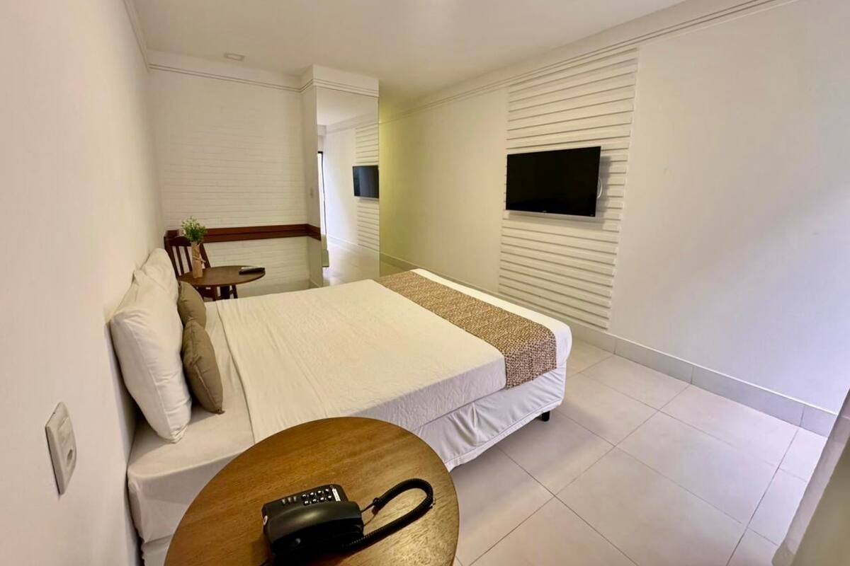 A bedroom with a bed, a bedside table with a phone, a small table with a plant, and a TV mounted on the wall.