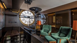 Een modern interieur met een grote wandklok, een spiltrap en groene bekleden stoelen.