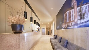 Een ontvangsthal met een receptiebalie, een grote kamerplant, een bank met kussens, en een muurschildering van een gebouw.