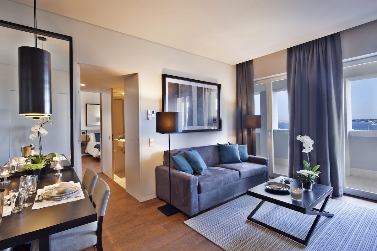 A modern hotel room with a dining area, a sofa, and a large window offering a view.