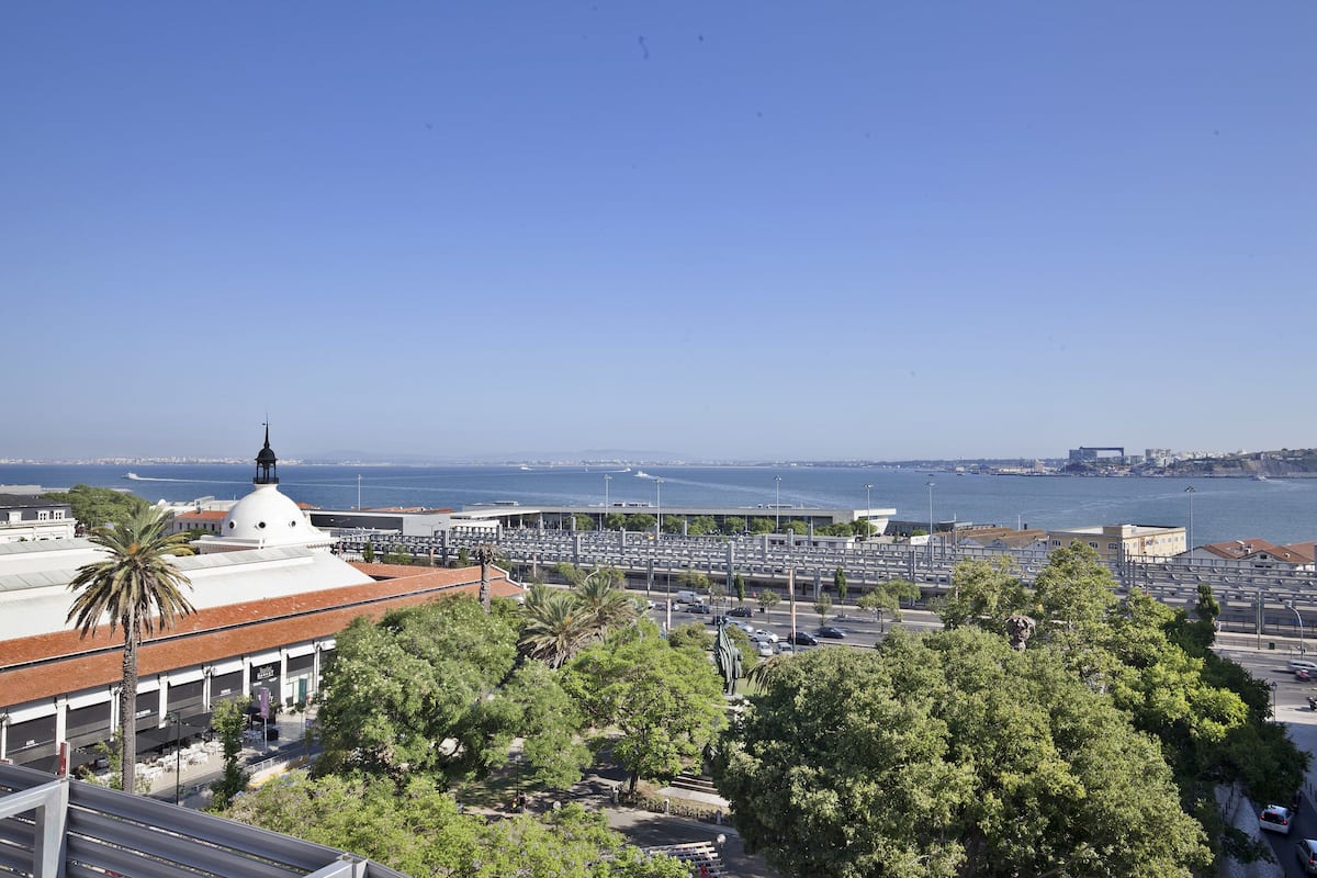 A coastal cityscape with a prominent white building with a dome and a statue, surrounded by lush green trees and a clear blue sky.