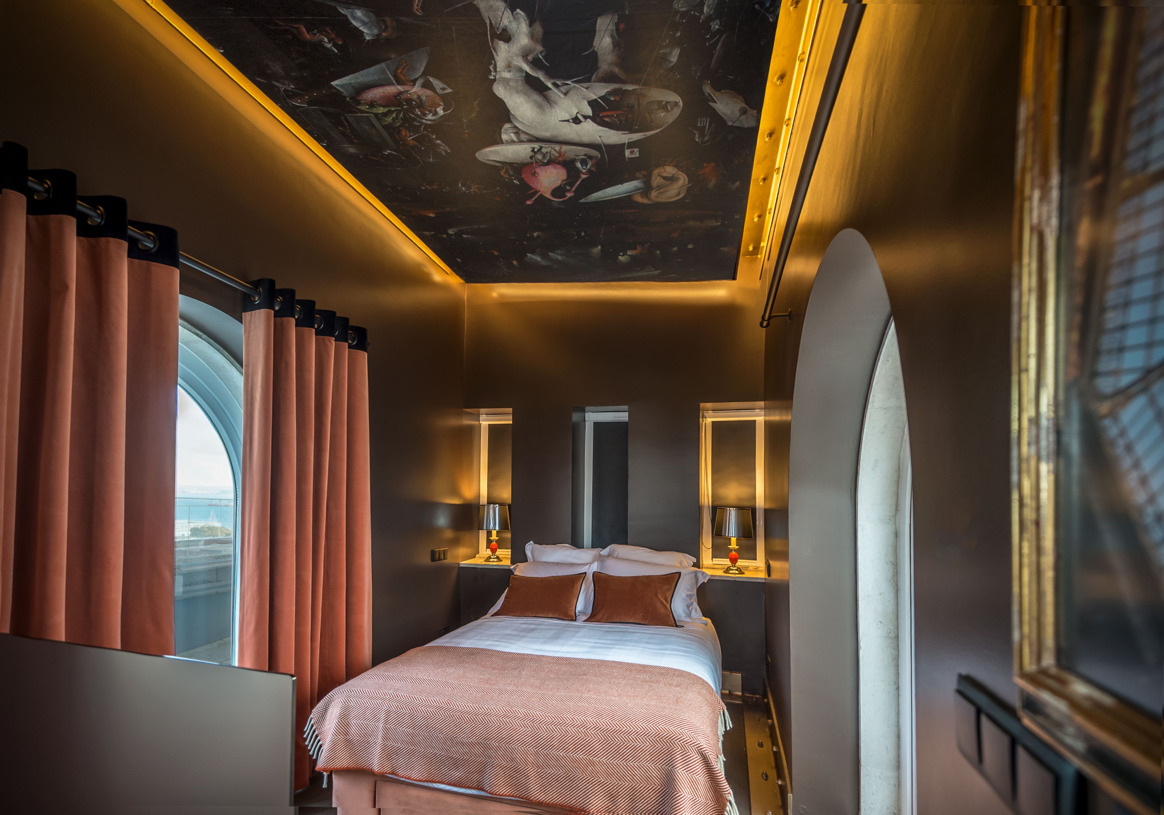 Une chambre d’hôtel moderne dotée d’un grand lit, de deux tables de chevet avec des lampes, d’une fresque au plafond et d’une grande fenêtre avec des rideaux.