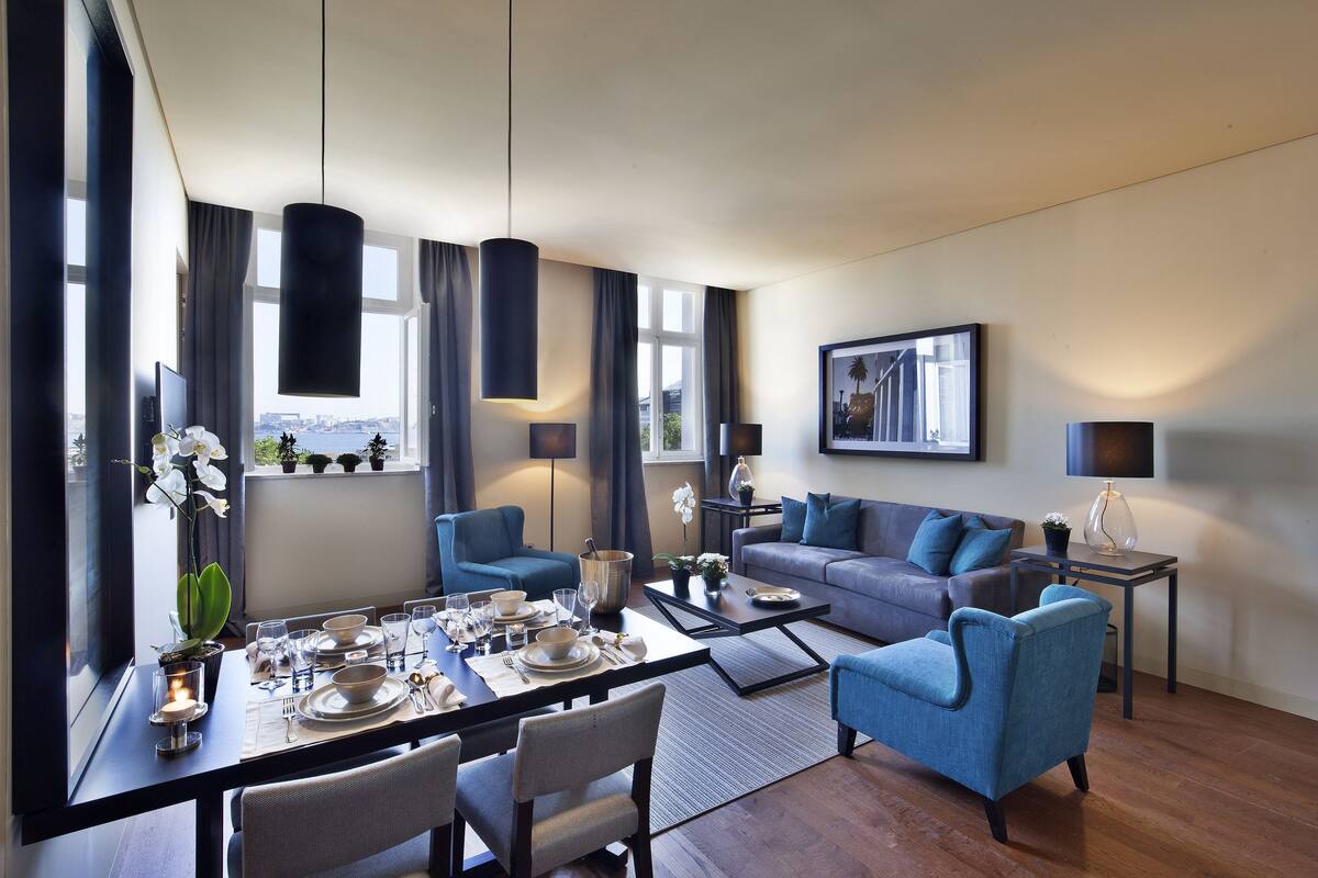 A modern living room with a dining area, blue furniture, and large windows offering a city view.