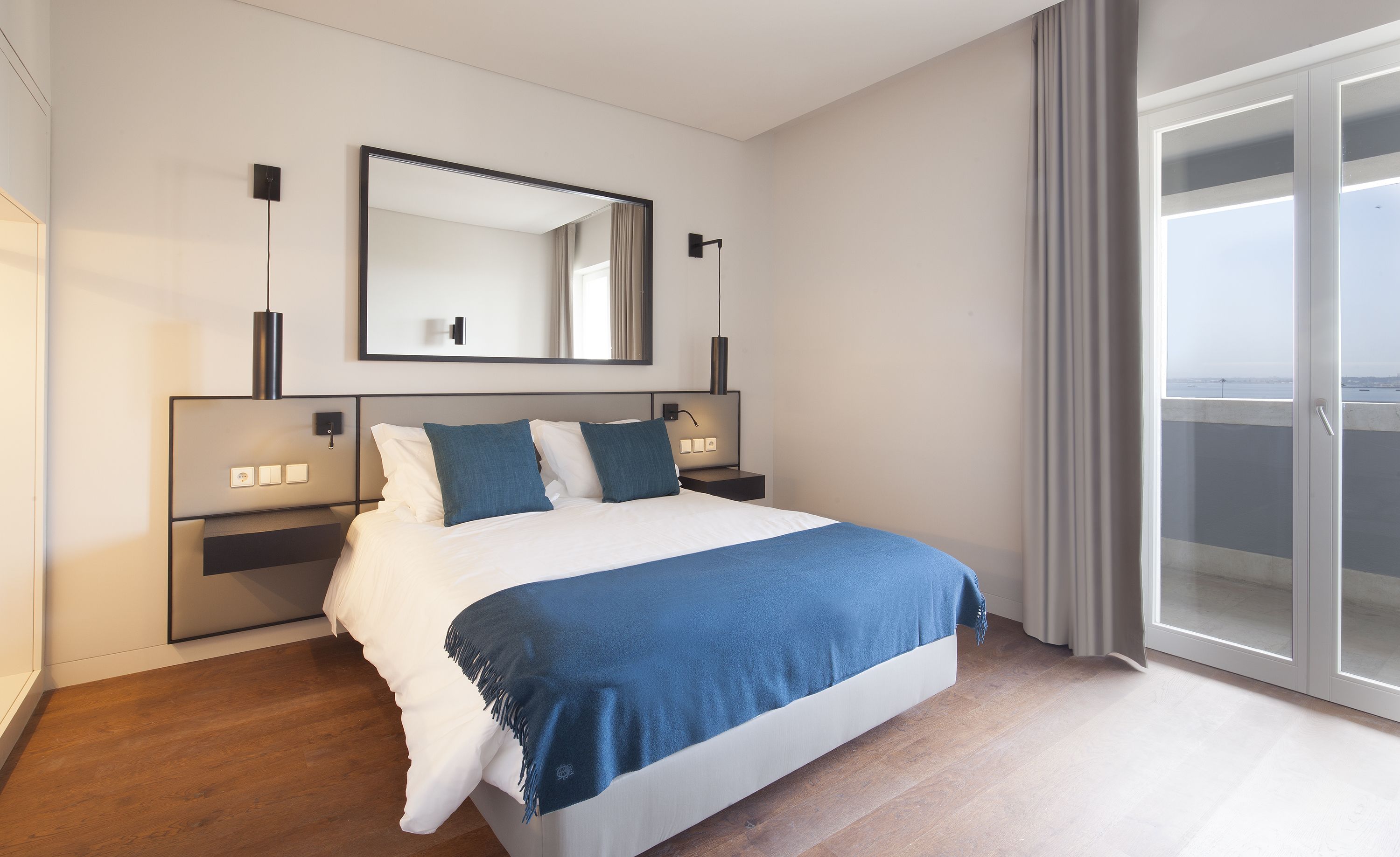 Una habitación de hotel moderna con una cama grande, una mesita de noche, un espejo y vista al mar desde la puerta del balcón.