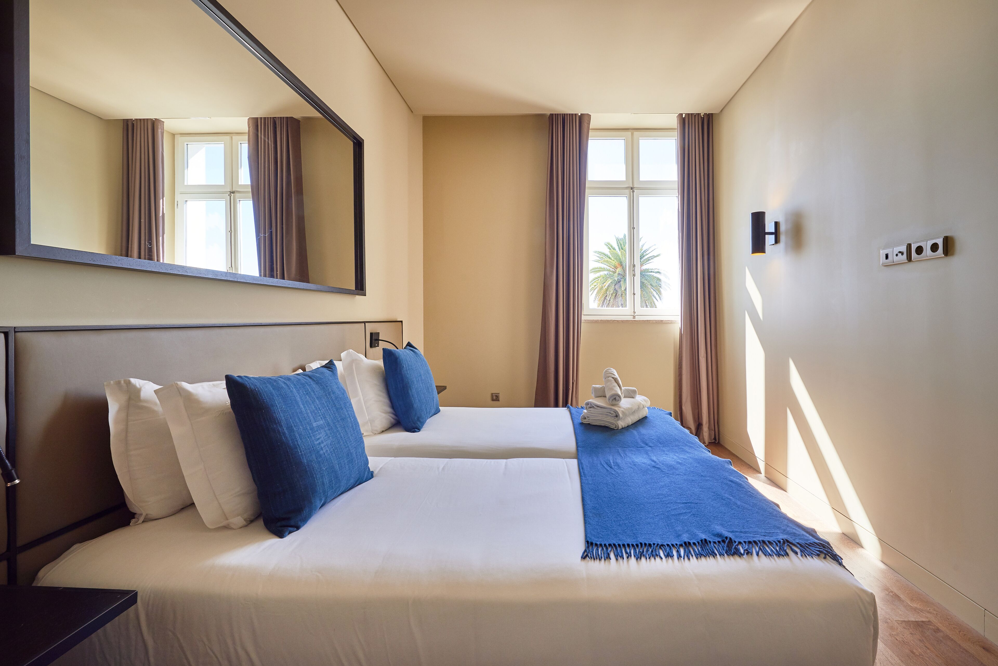 Een hotelkamer met twee bedden, een grote spiegel, een raam met gordijnen en een wandlamp.