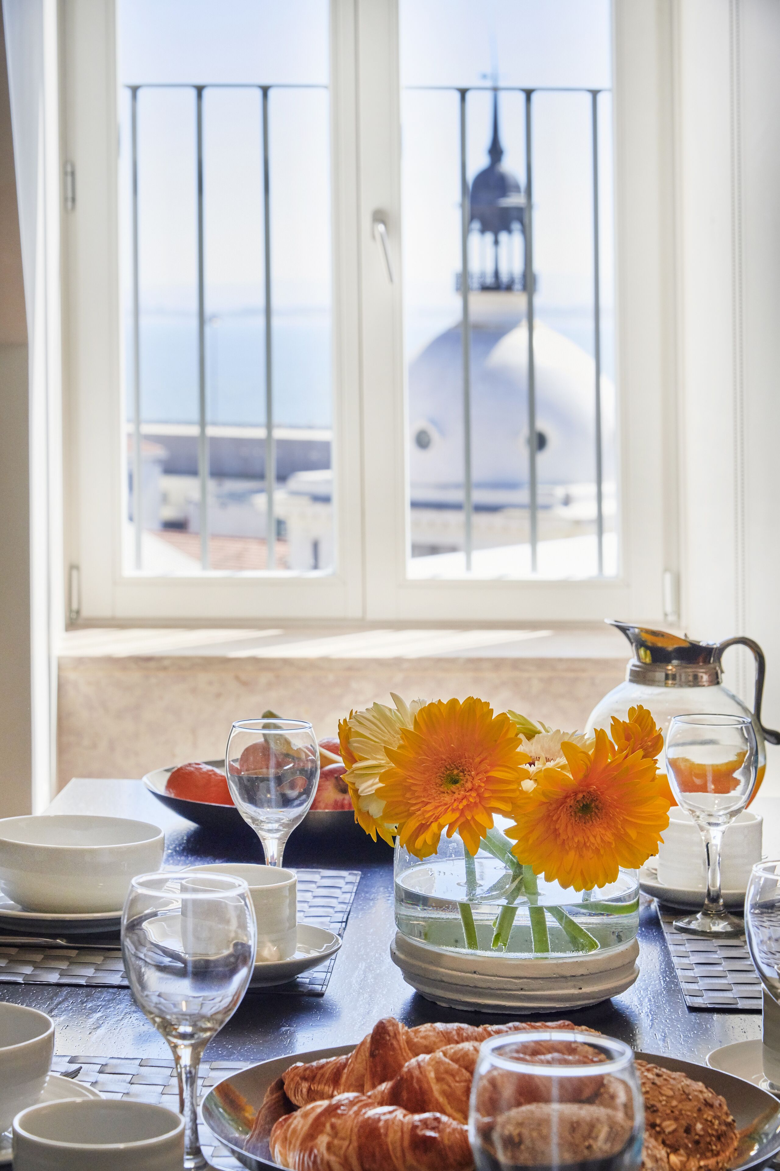A breakfast table with croissants, a vase with flowers, and a view of a building through a window.
