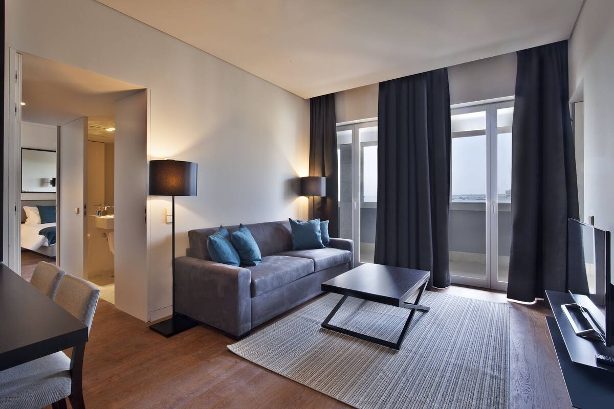 A modern hotel room with a sofa, a dining table, a TV, and a large window with curtains.