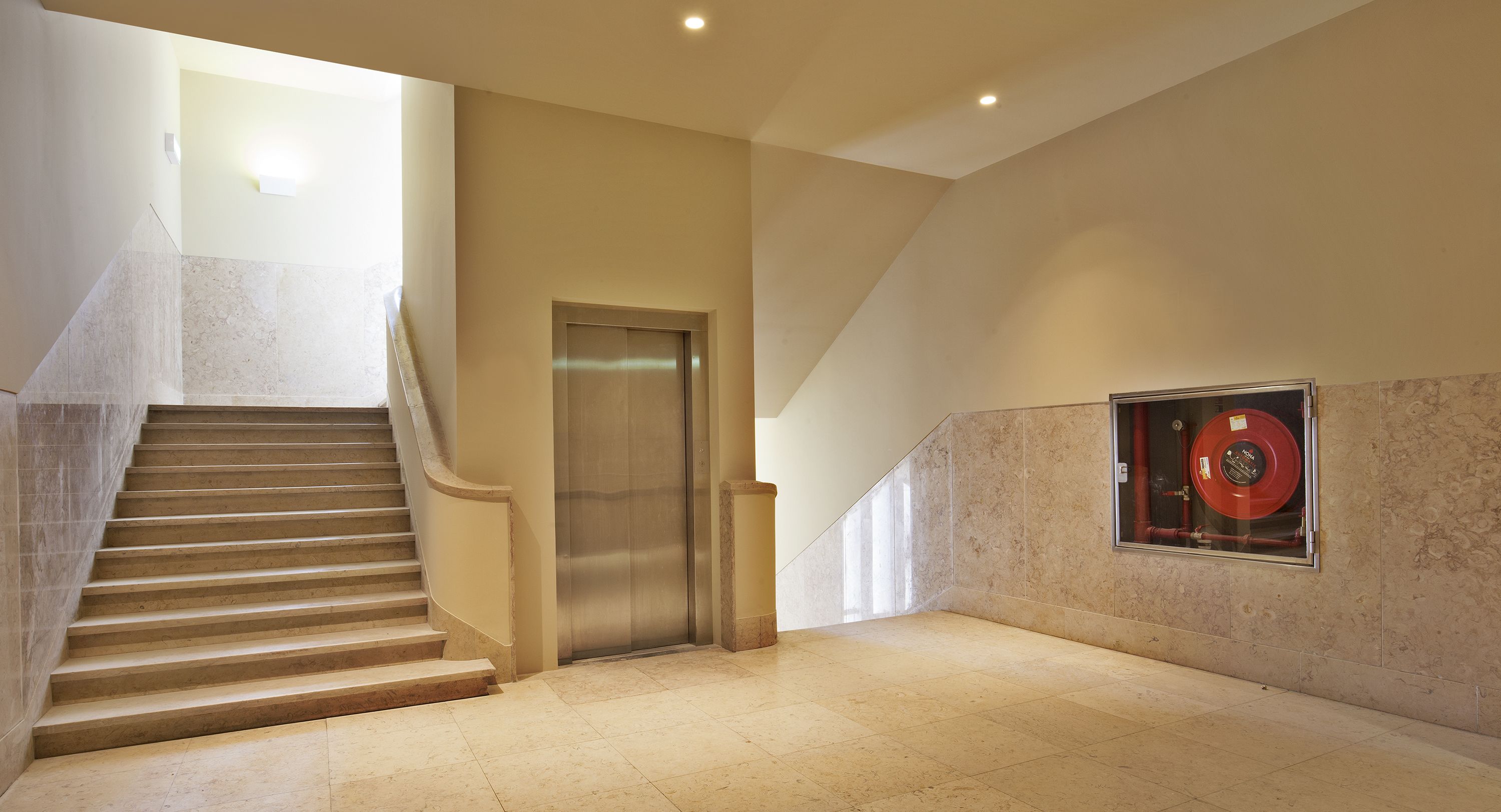 Un interior moderno con una escalera de mármol, un ascensor y un objeto circular rojo sujeto a la pared.