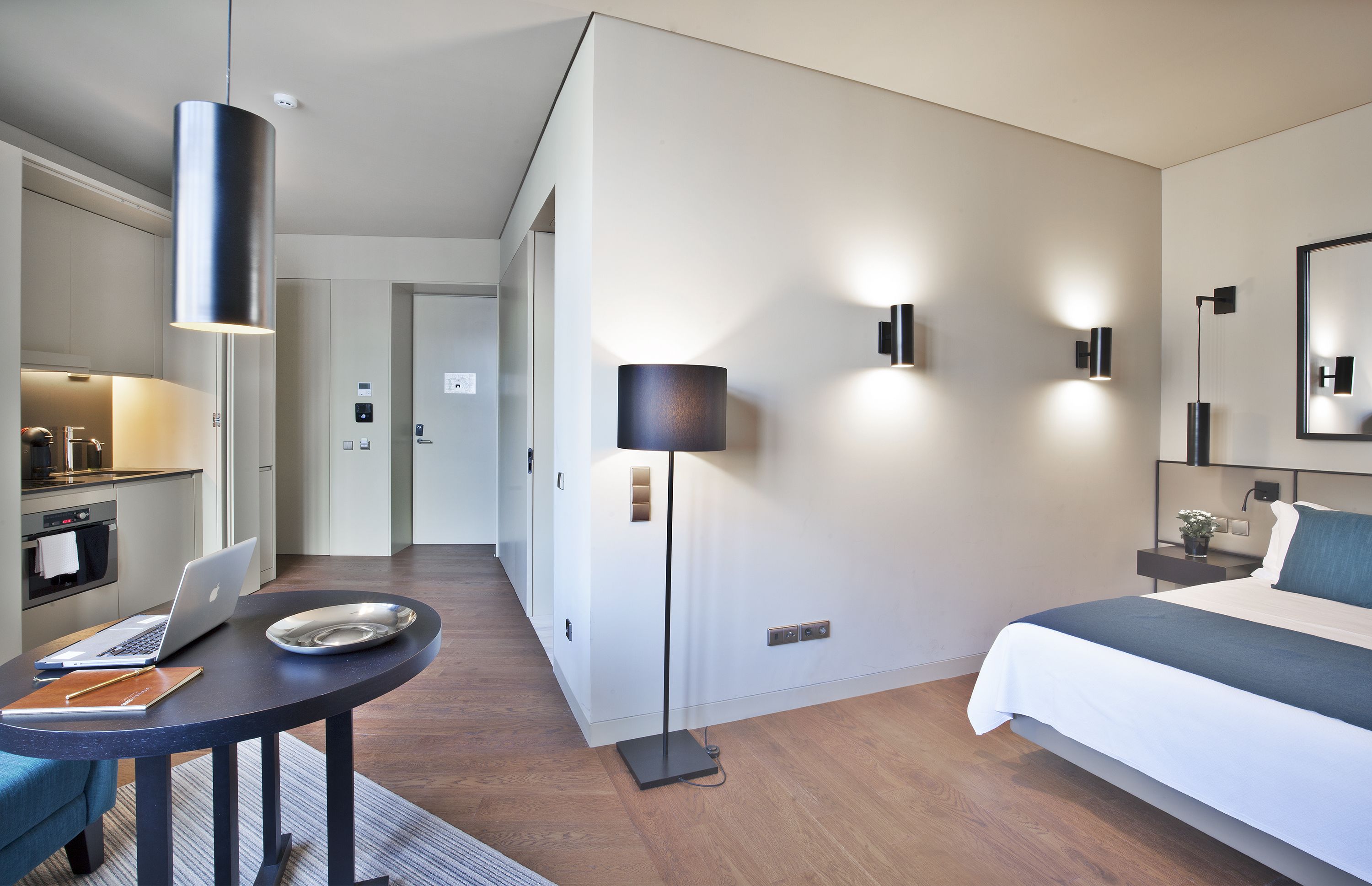 Ein modernes Hotelzimmer mit einem Bett, einem Schreibtisch mit einem Laptop, einer Lampe und einem kleinen Tisch mit Notizbuch und Stift.