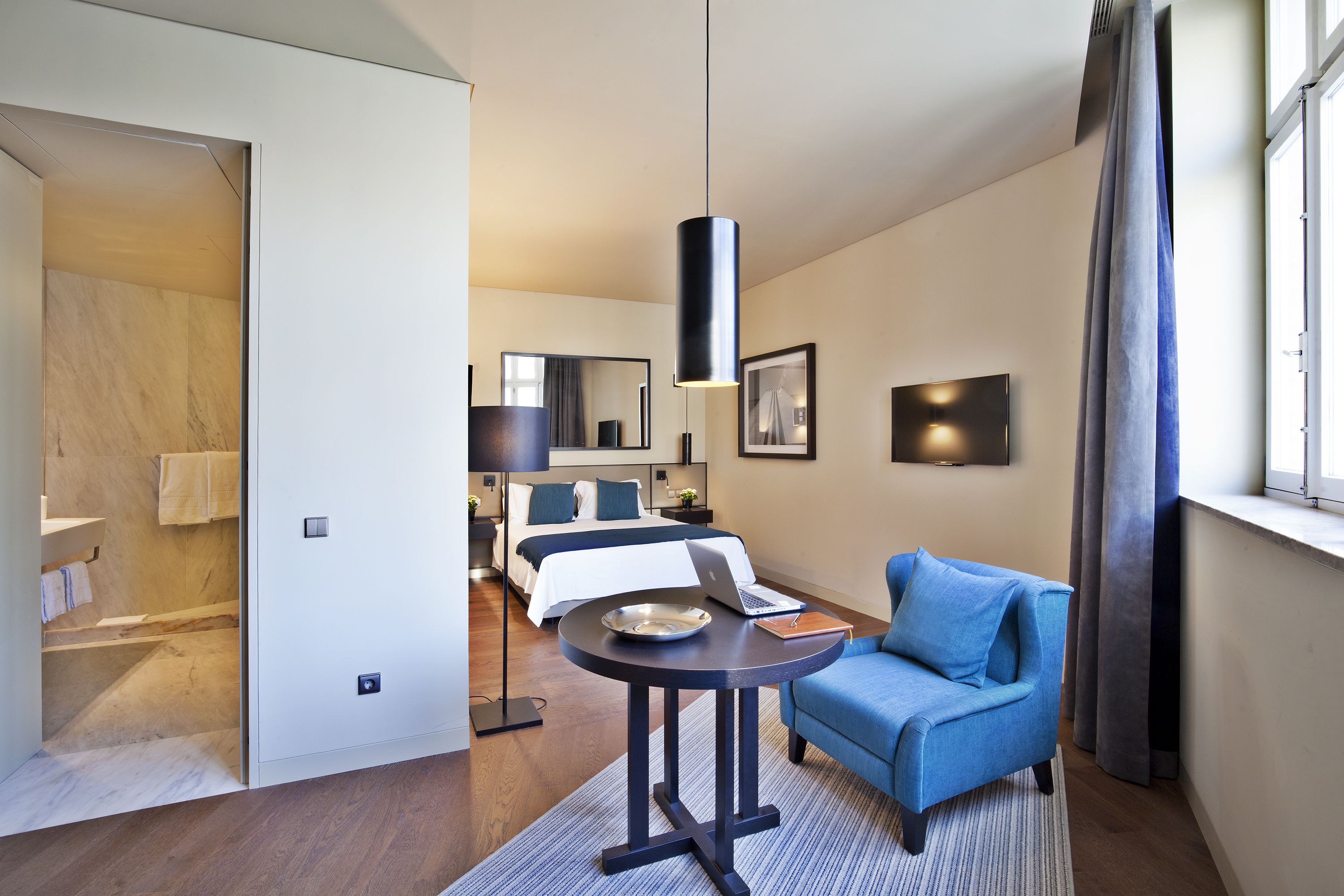 A modern hotel room with a bed, a blue sofa, a round table, and a large window with curtains.