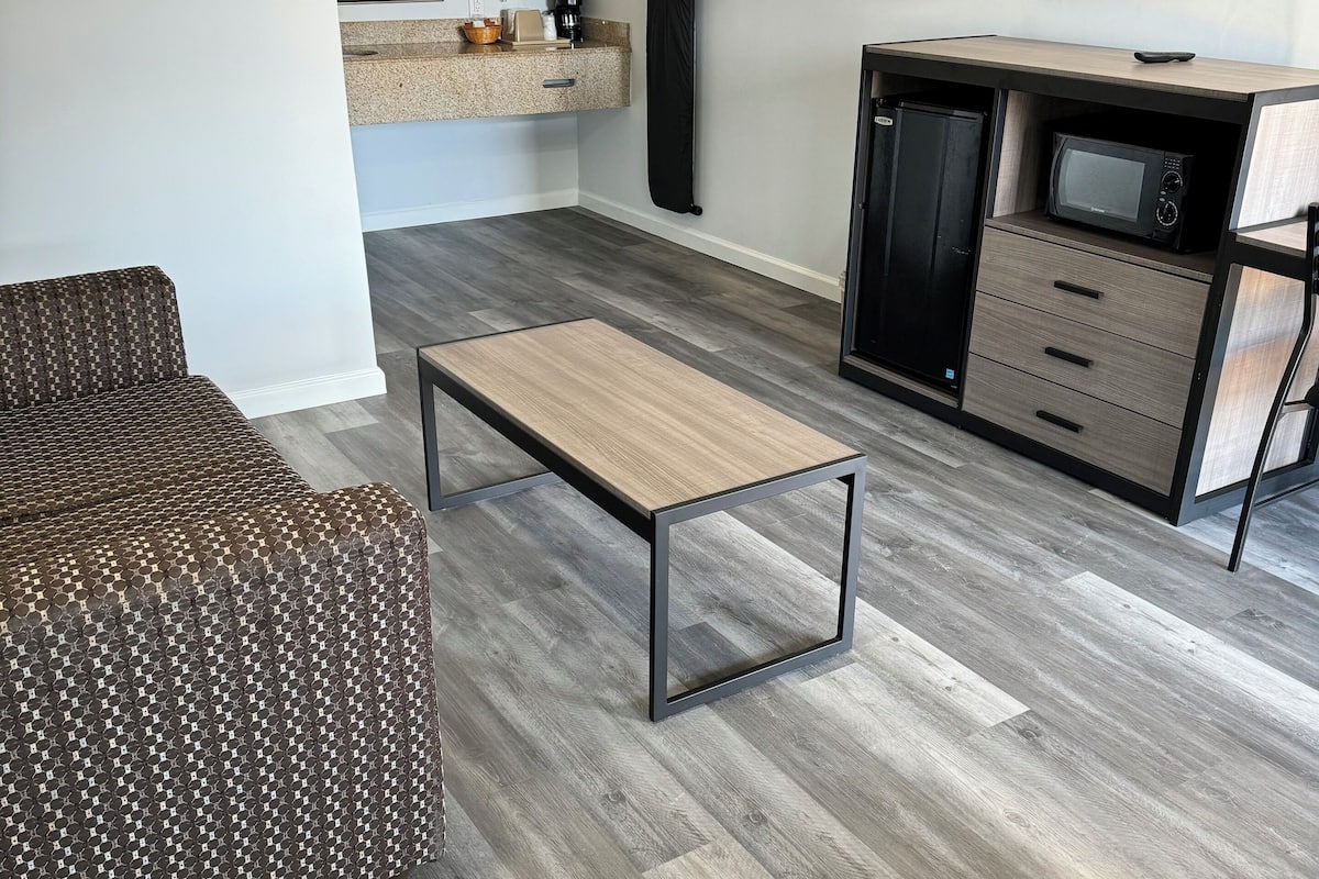 Uma sala de estar moderna com um sofá estampado, uma mesa de centro de madeira e uma área de cozinha compacta com micro-ondas e armários.