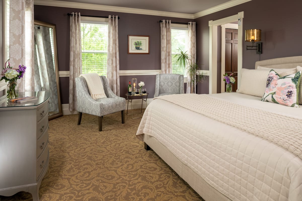A bedroom with a large bed, two armchairs, a small table with a vase of flowers, and a mirror.