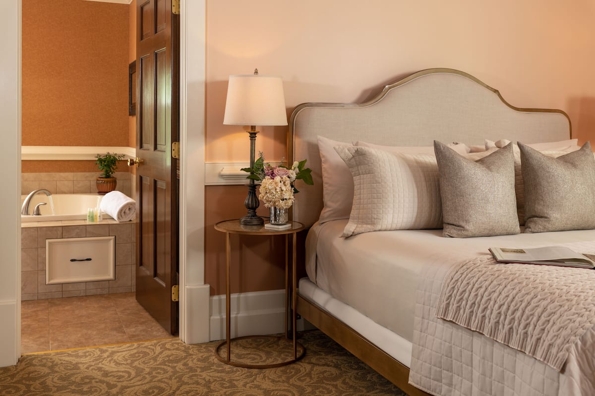 A hotel room with a bed, a bedside table with a lamp and flowers, a bathroom with a sink and a towel rack.