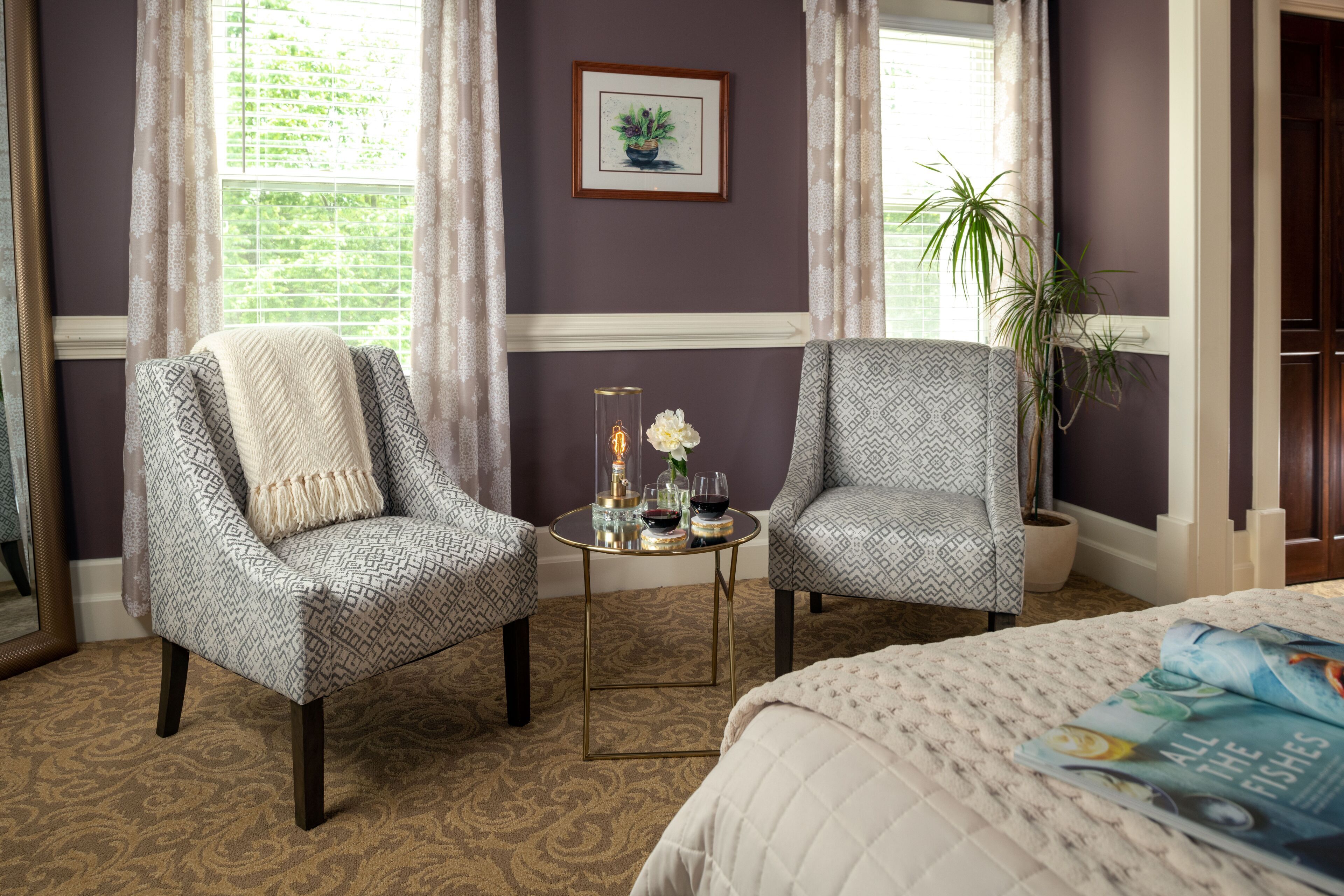 Une pièce comprenant deux fauteuils à motifs, une petite table ronde avec un vase et des verres à vin, un lit avec une courtepointe et une plante en pot.