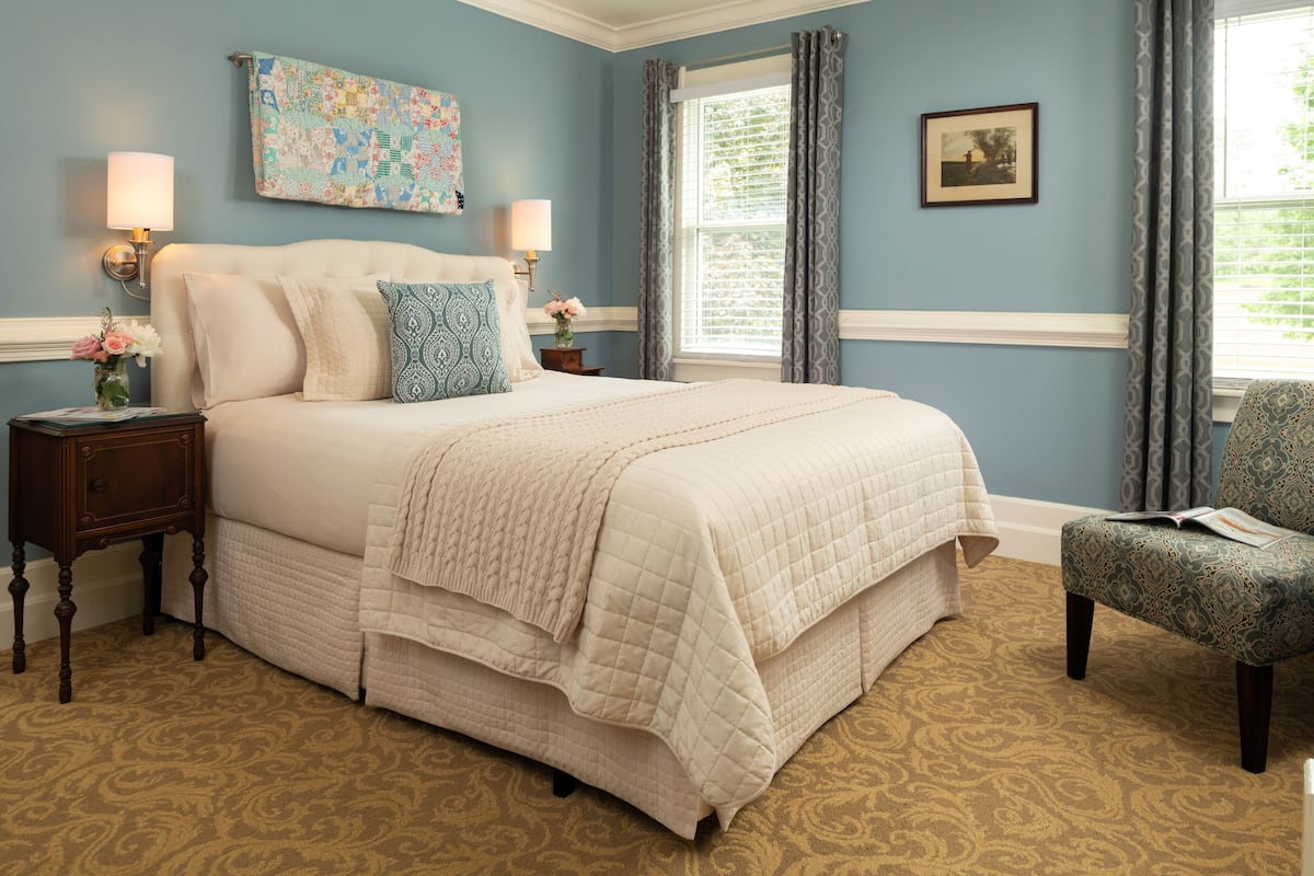 A bedroom with a large bed, two bedside tables, a chair, a window with curtains, and a wall with a picture and a quilt.