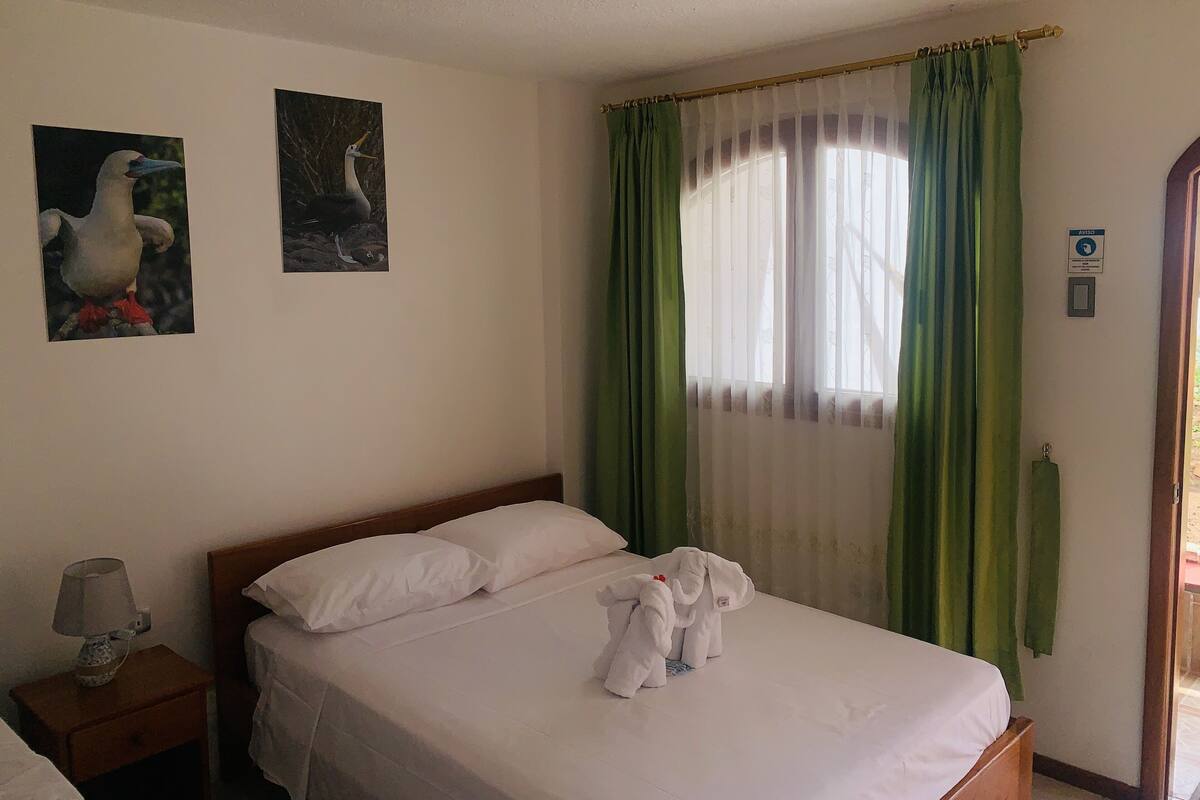 Hotellihuone, jossa on sänky, yöpöytä lampun kanssa, kaksi seinälle ripustettua taulua, ikkuna vihreillä verhoilla ja kyltti, jossa lukee AVEJO.