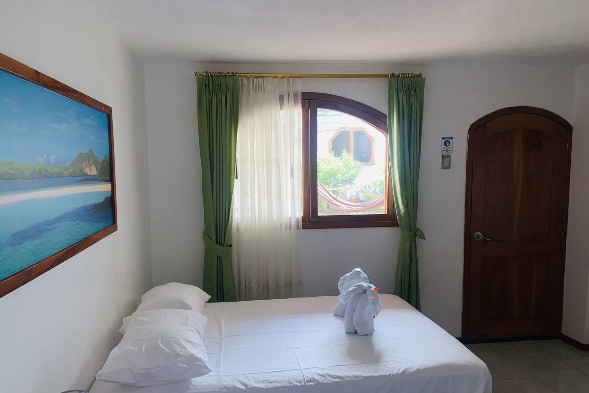 Hotellihuone, jossa on sänky, ikkuna verhoilla, ovi ja taulu seinällä.