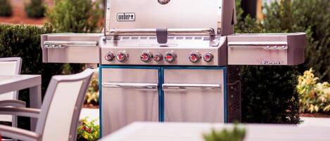 Ein Weber Summit Gasgrill mit vier Brennern, einem Thermometer und einer Bedieneinheit, draußen aufgestellt, mit einem Tisch und Stühlen in der Nähe.