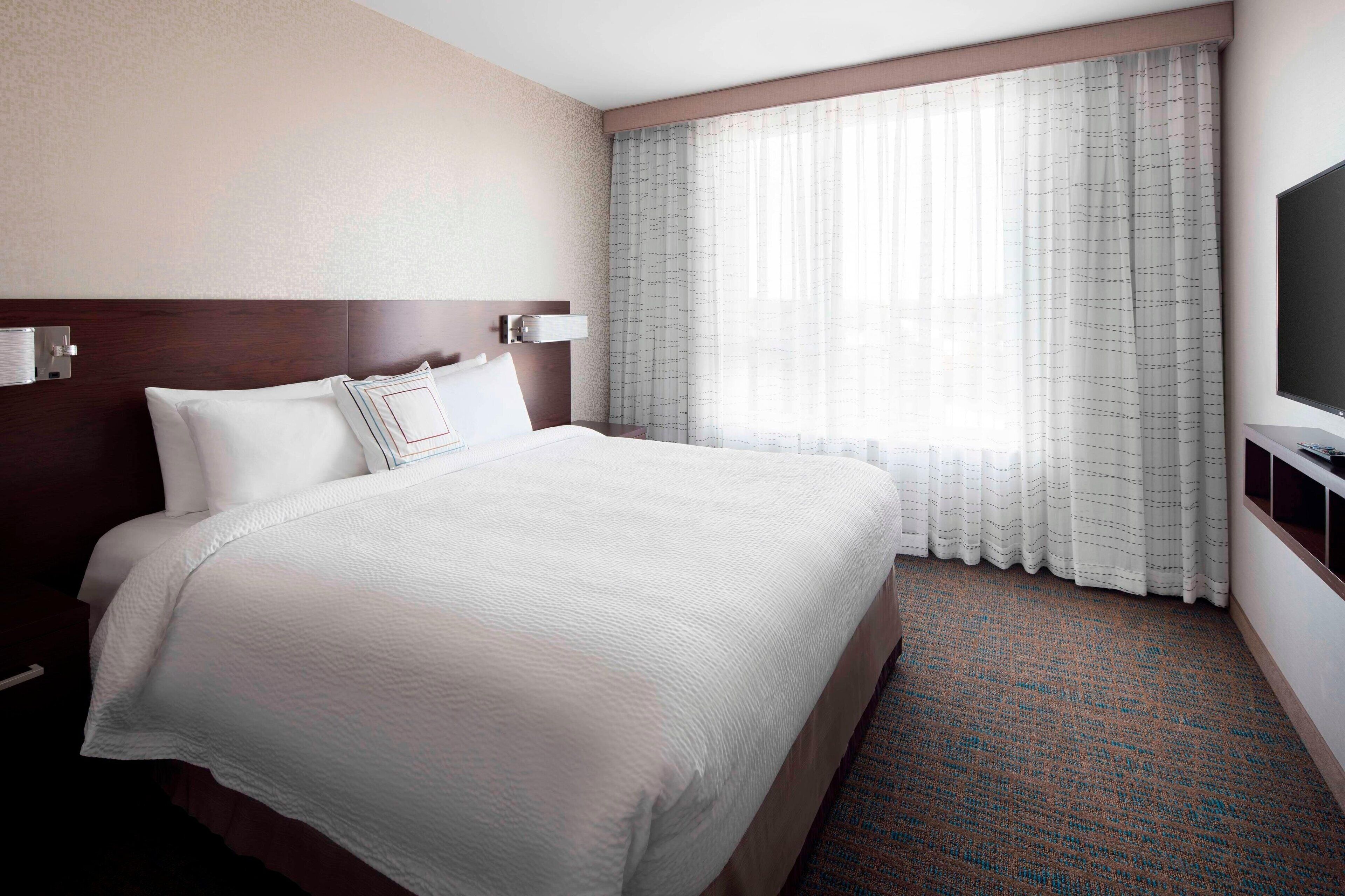A hotel room with a large bed, a flat-screen TV, a window with sheer curtains, and a carpeted floor.