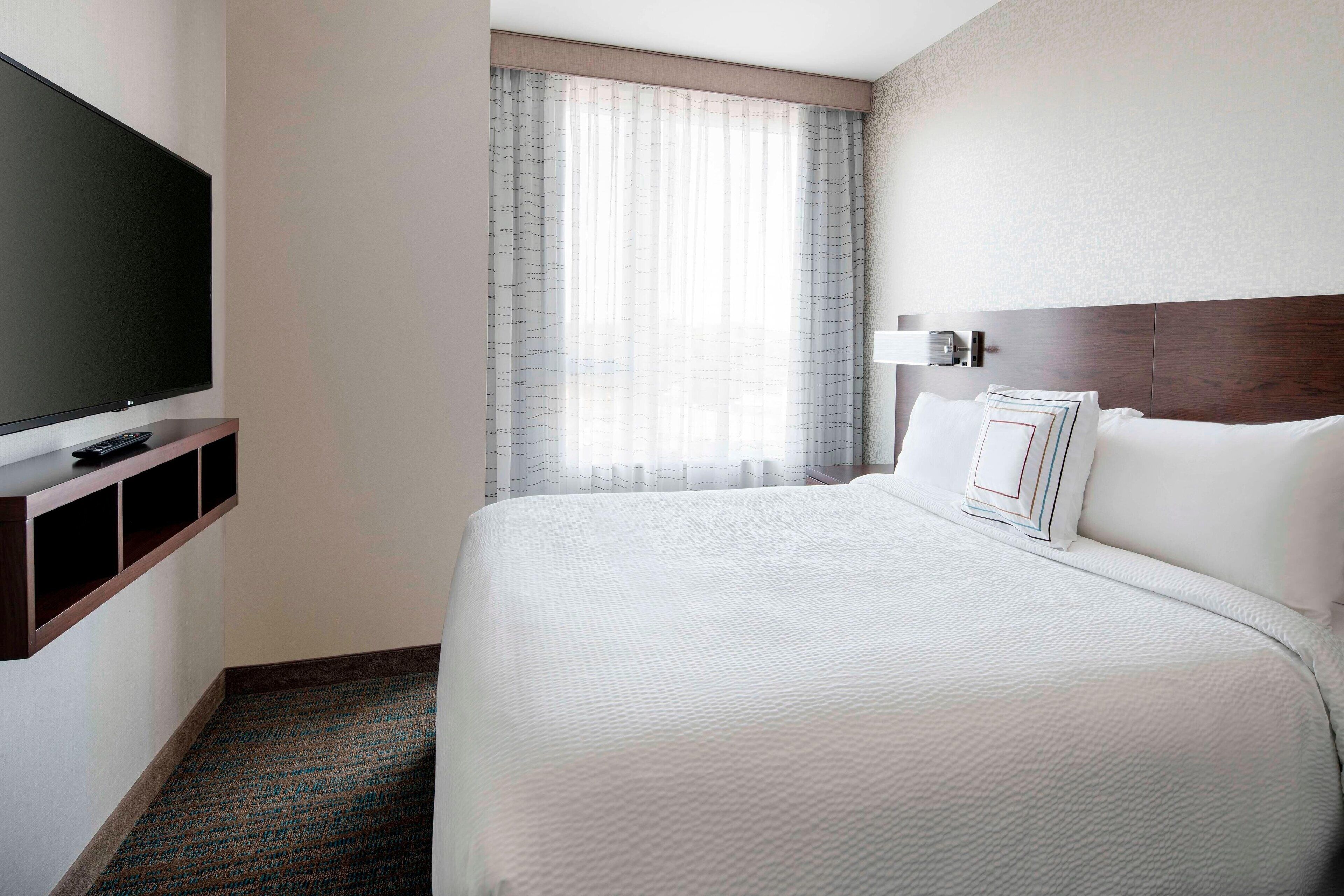 A hotel room with a large bed, a flat-screen TV on a wooden shelf, a window with sheer curtains, and a wall-mounted lamp.