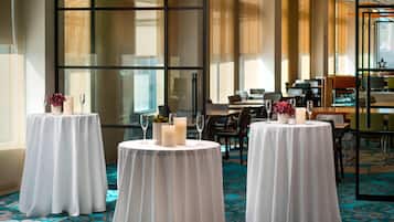 Une salle à manger d’hôtel avec des tables rondes disposées pour deux personnes, chacune agrémentée d’une nappe blanche, de bougies et de compositions florales. De grandes fenêtres laissent entrer la lumière naturelle et illuminent l’espace.