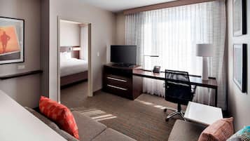 A hotel room with a bed, a desk with a TV, a chair, a sofa, and a window with curtains.