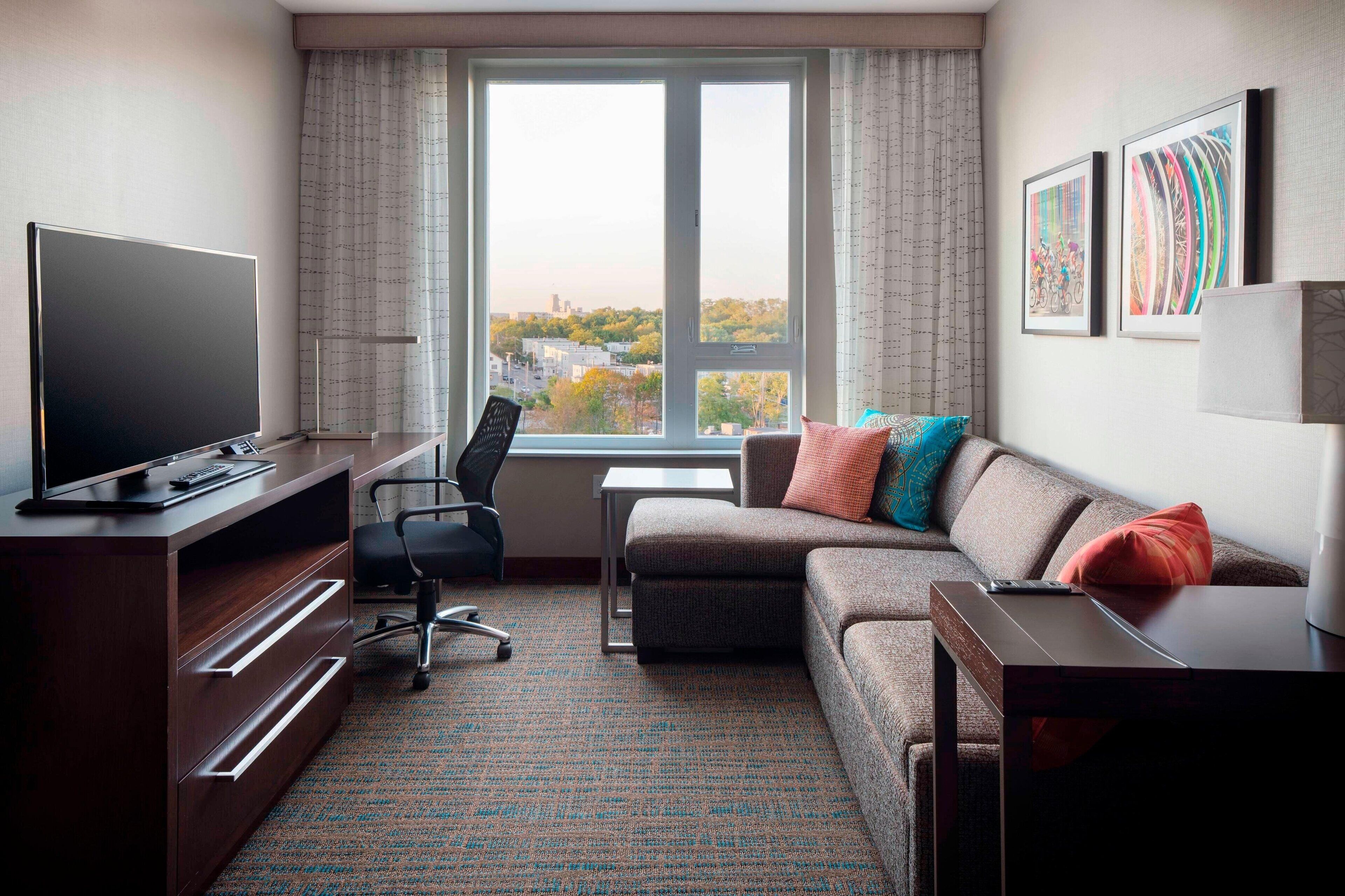 A modern hotel room with a large window, a flat-screen TV, a sofa, a desk, and a chair.