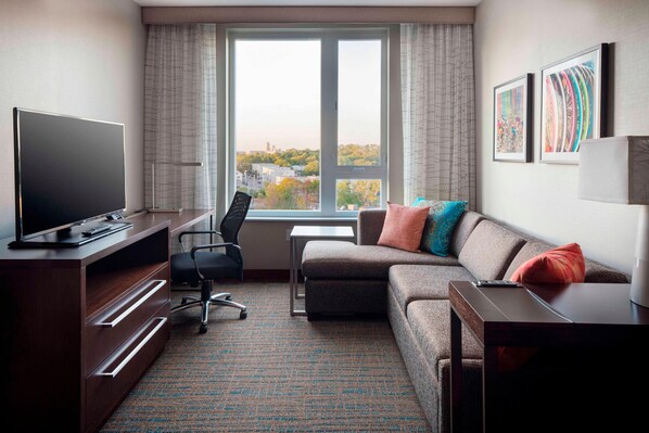 A modern hotel room with a large window, a flat-screen TV, a sofa, a desk, and a chair.