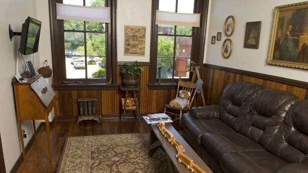 A cozy living room with a leather sofa, a wooden coffee table with a model train, a rocking chair, and a large window with a view of trees and parked cars. - The Tinder Guest House (Ashland)